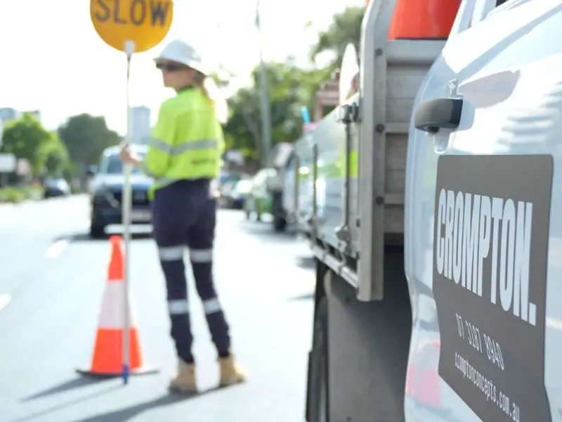 Traffic control worker holding slow sign beside Crompton vehicle and roadside safety cones